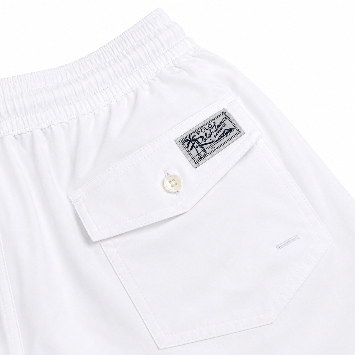 Close-up of white shorts showing the back waistband, a flap pocket with a button, and a rectangular embroidered brand patch reading 'Polo Ralph Lauren'.