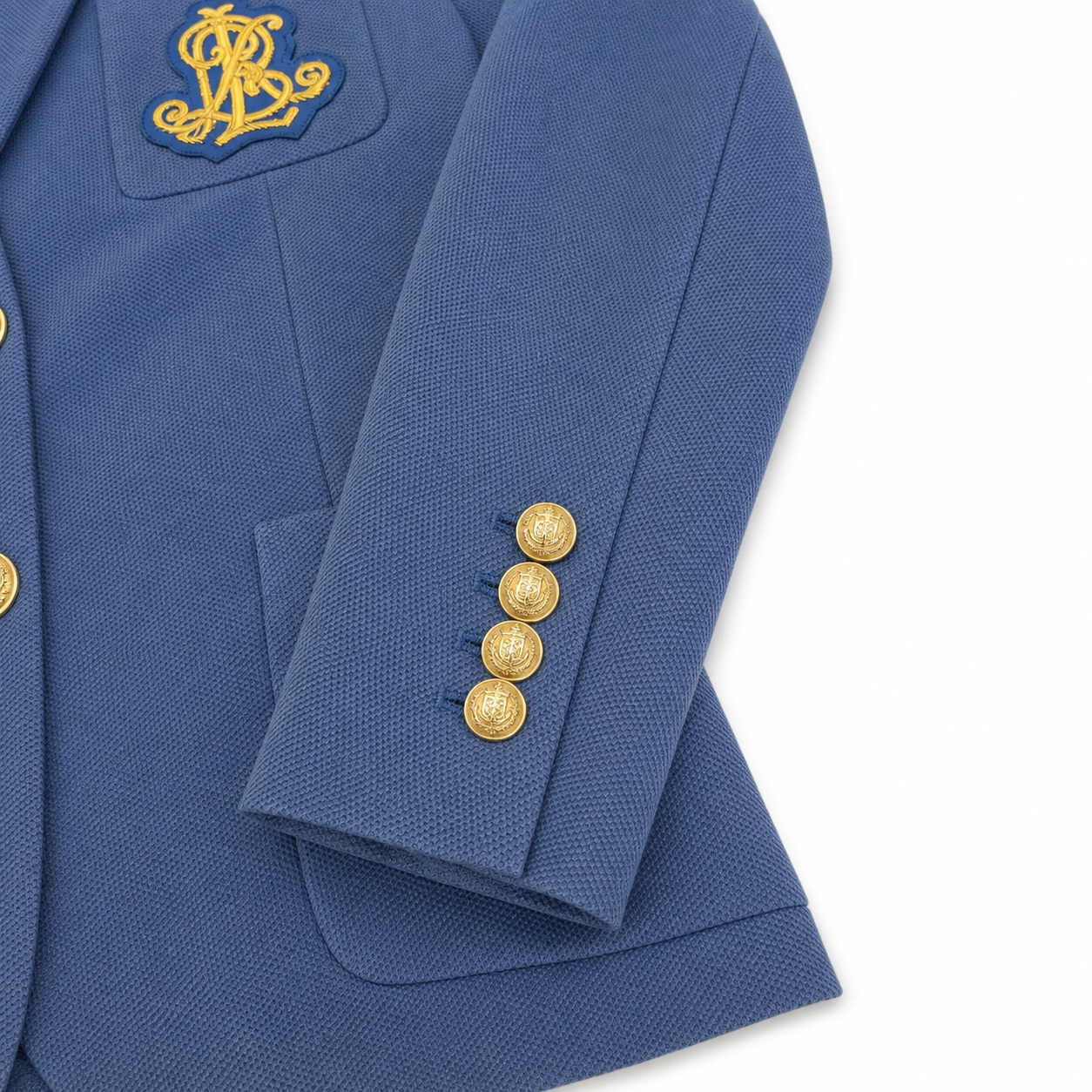 Close-up of a blue blazer with gold embroidered monogram patch on the chest and four gold buttons on the cuff, shown on a white background.