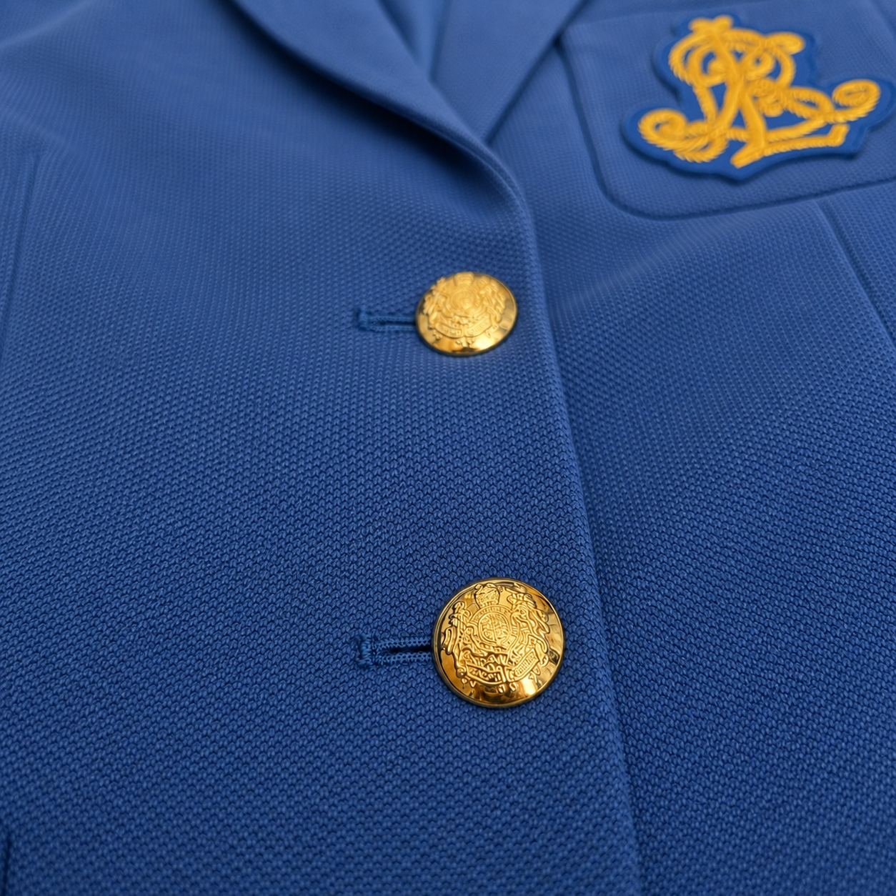 Close-up of a royal blue textured jacket with two prominent gold buttons and a gold embroidered crest patch on the chest pocket.