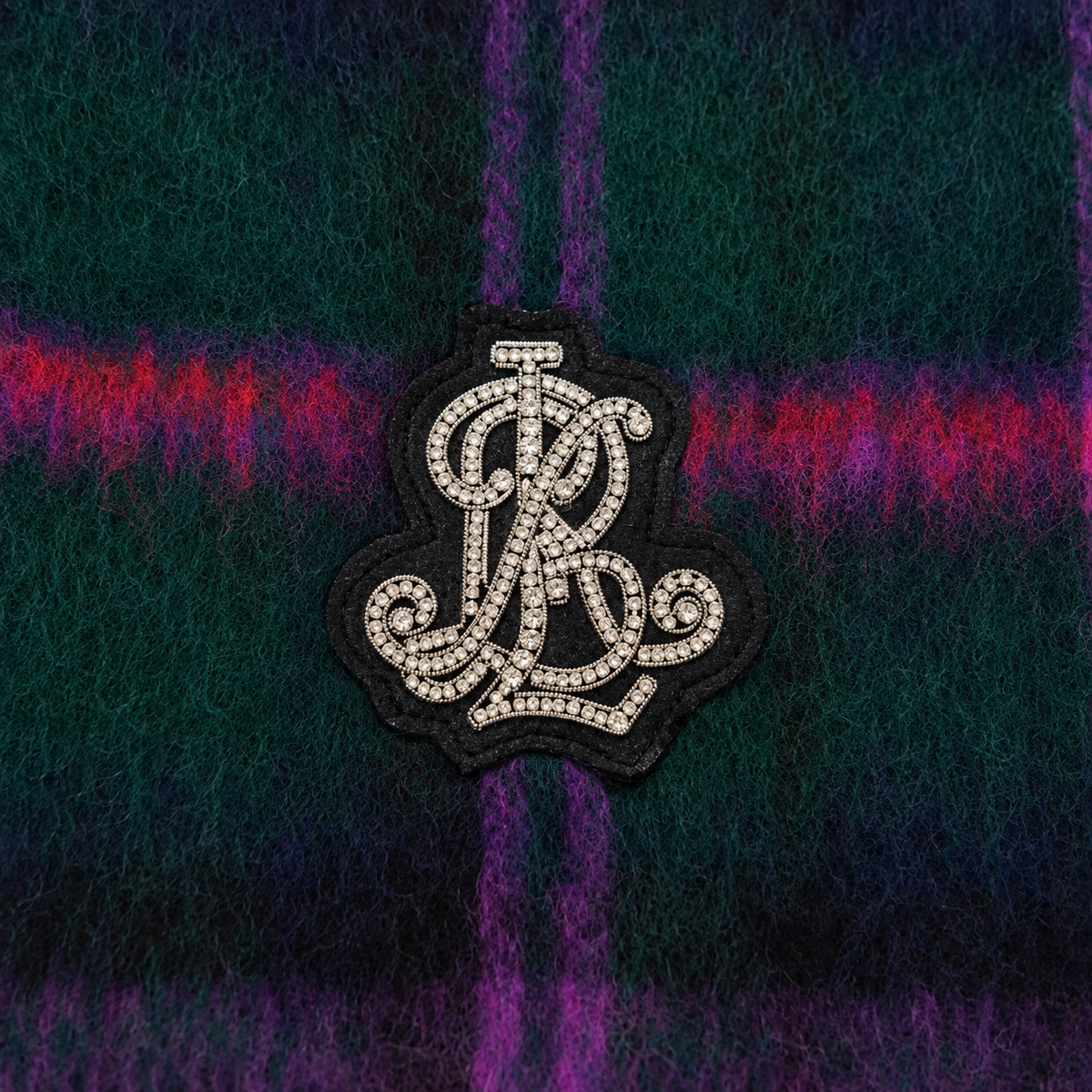 A close-up of a black embroidered patch with a white monogram logo, placed on a dark green and purple plaid fabric background.