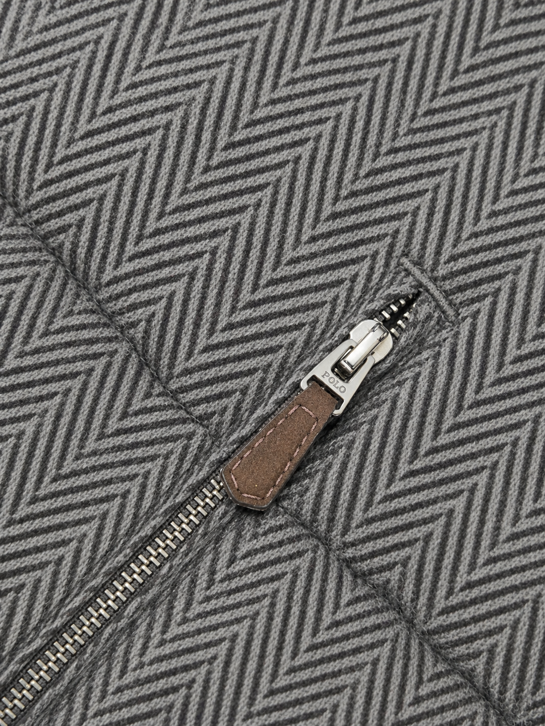 Close-up of a gray herringbone-patterned fabric with a silver zipper and a brown leather pull tab.
