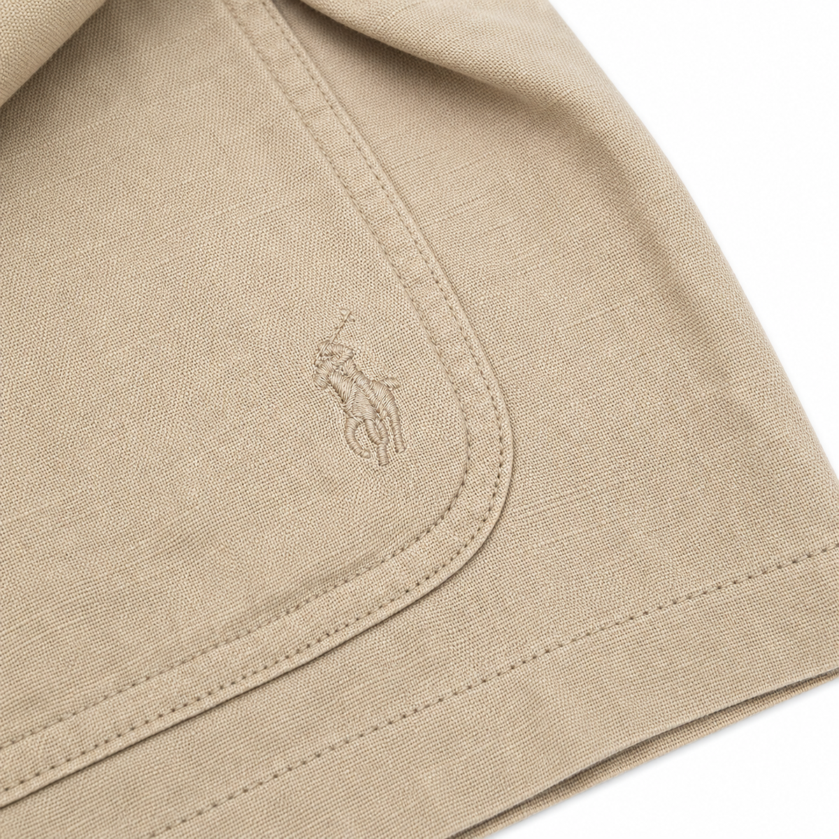 Close-up of a beige woven textile with a curved stitched edge and a small tonal embroidered polo player logo, shown on a white background.