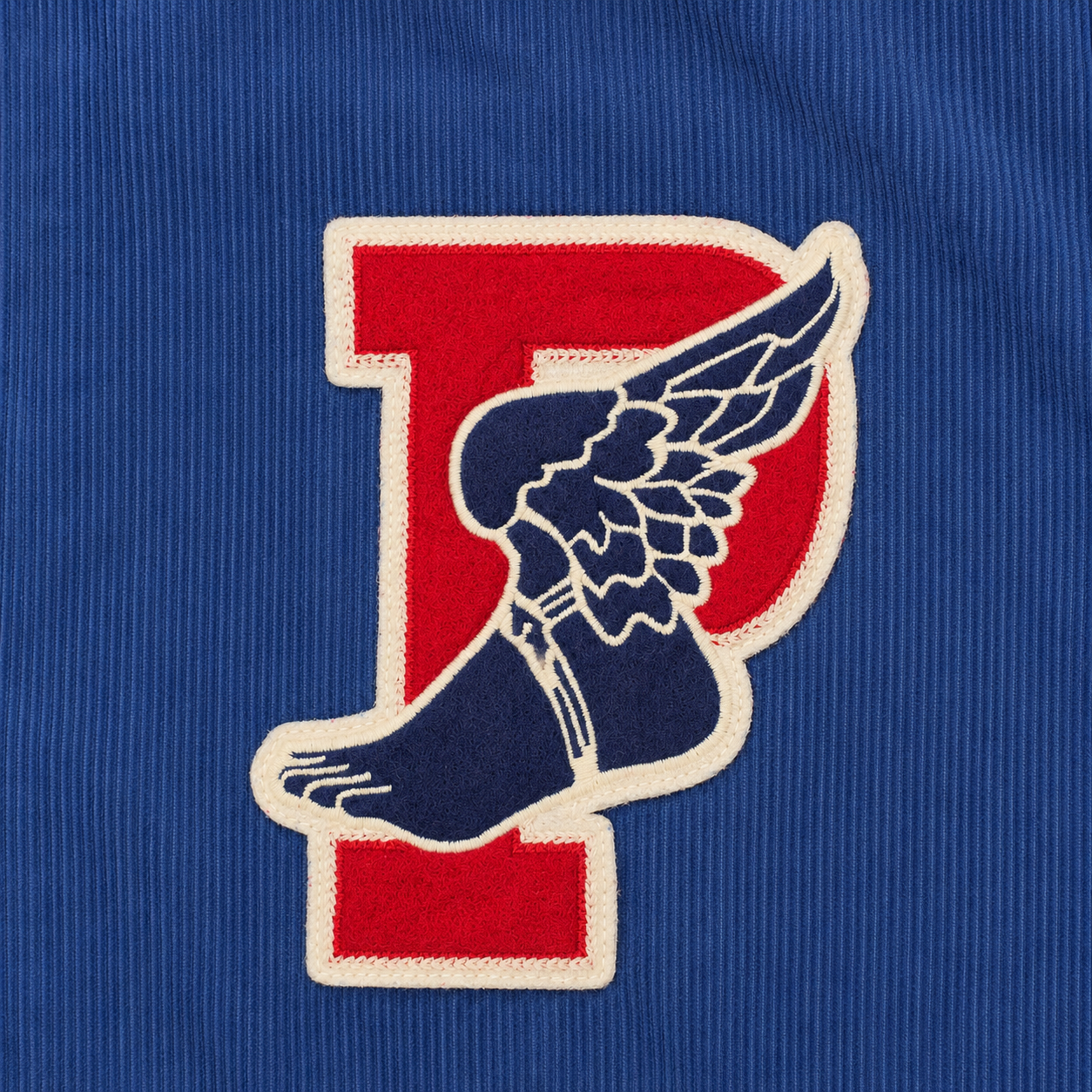 A close-up of a blue ribbed fabric featuring a large embroidered letter P logo with a winged foot design in red, navy blue, and cream.