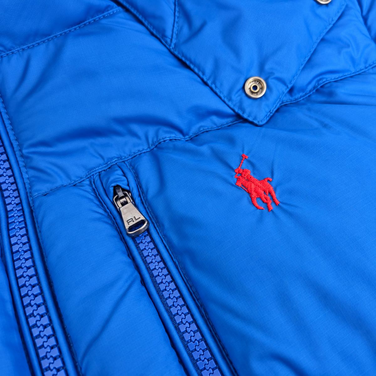 Close-up of a bright blue puffer jacket showing the red embroidered Polo pony logo, a blue zipper with a silver pull, and snap buttons near the collar.
