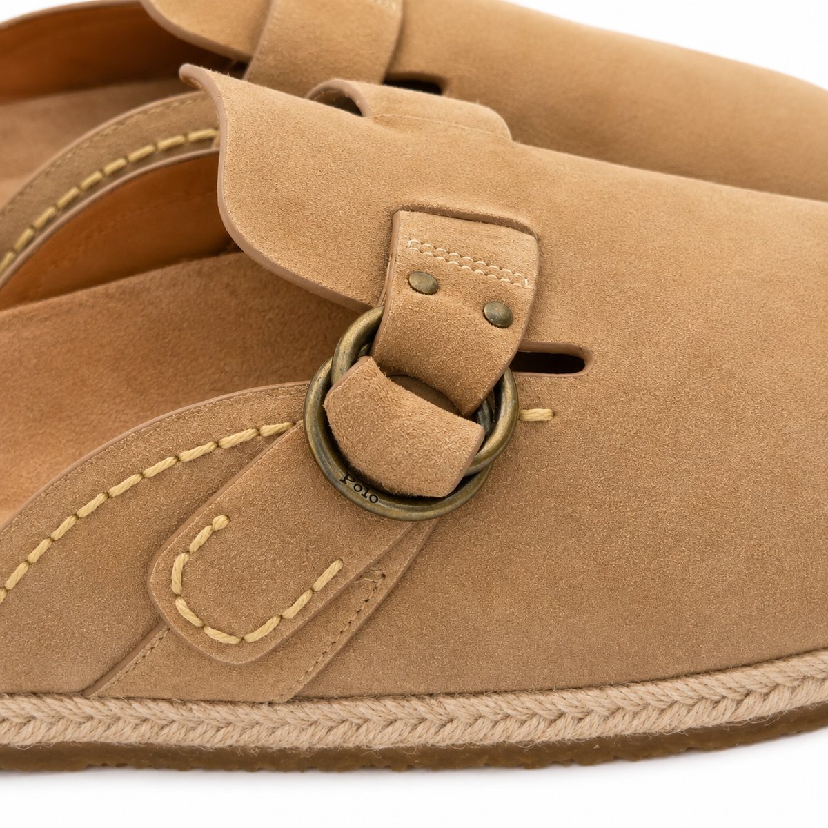 Close-up of a tan suede clog-style shoe with a strap, metal ring hardware, contrast stitching, and a braided jute sole edge on a white background.